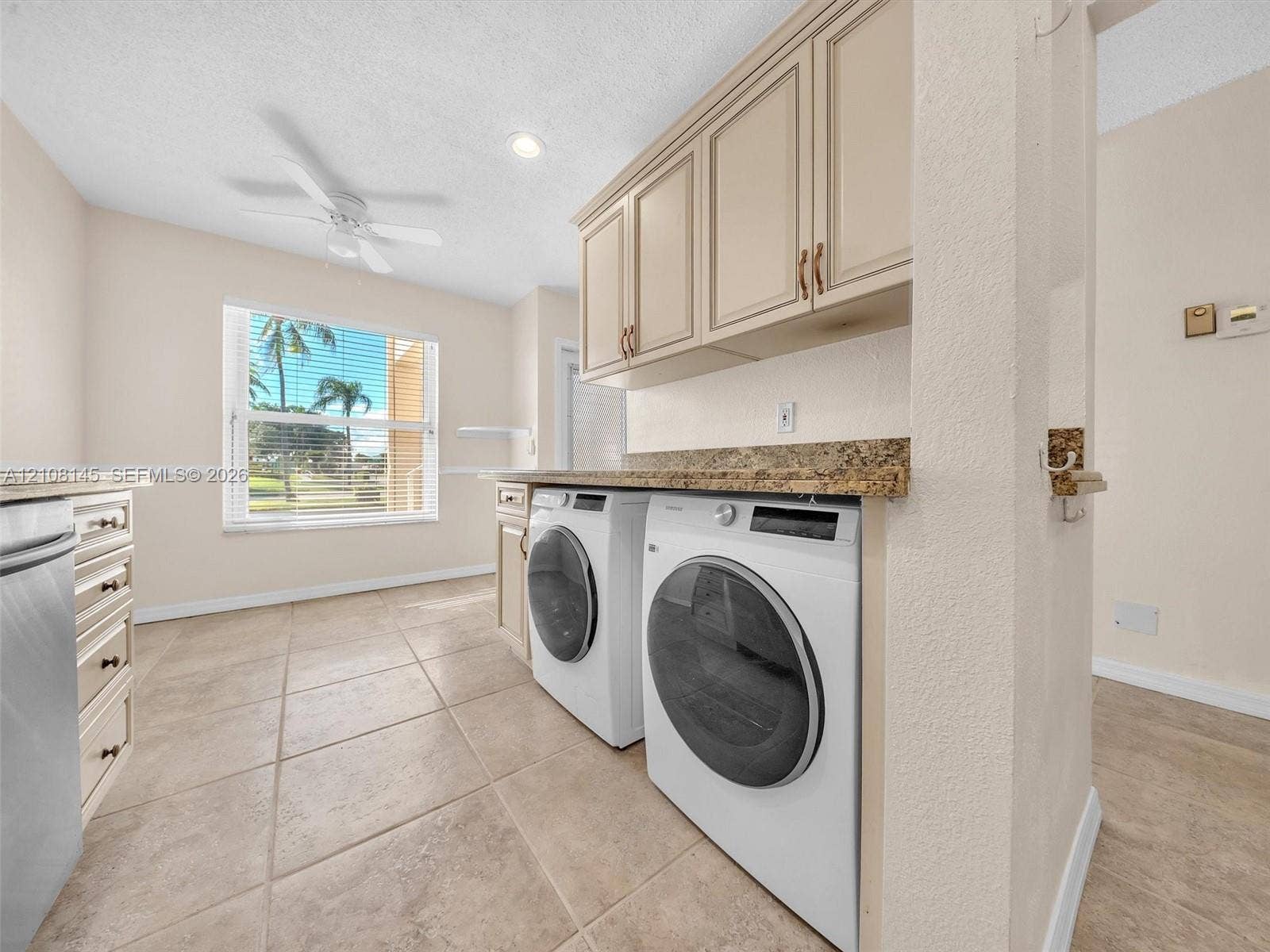 1020 Citrus Way, #101, Delray Beach, FL 33445 Laundry area featuring stone-finish countertops, wall-mounted cabinetry, tile flooring, a ceiling fan, and a window with exterior scenery