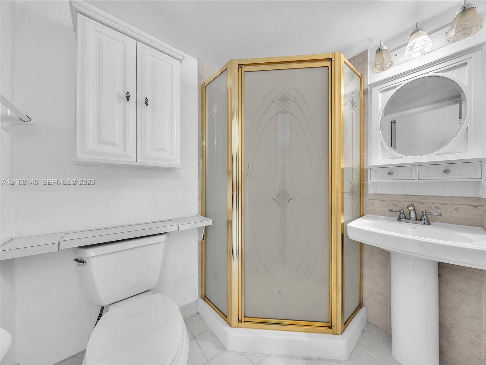 1020 Citrus Way, #101, Delray Beach, FL 33445 Corner shower with brass-finish frame and etched glass doors, white pedestal sink with dual-handle faucet, circular wall-mounted mirror, white shaker-style wall cabinetry, and tile flooring