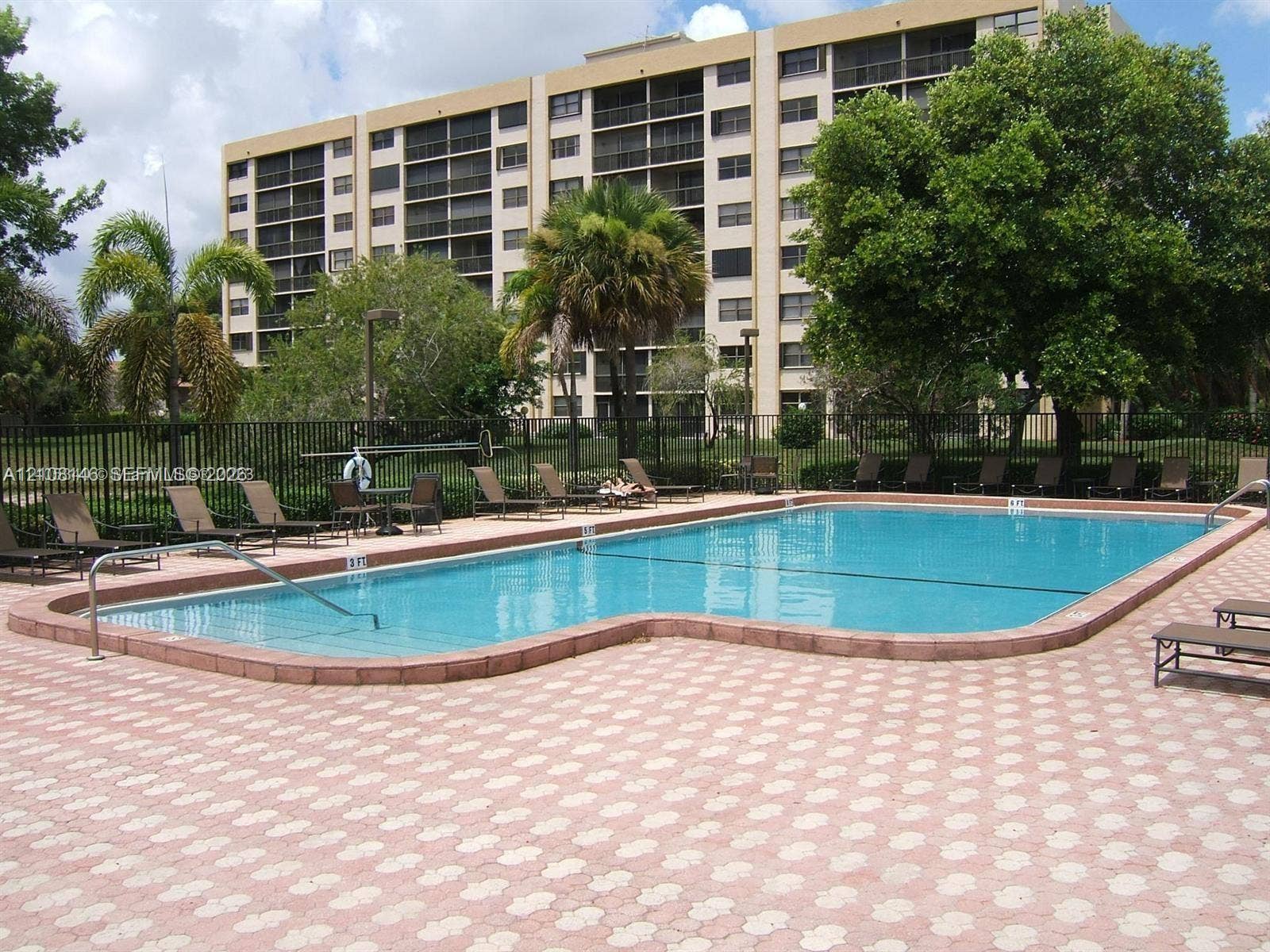 1258 S Military Trail, #813, Deerfield Beach, Deerfield Beach, FL 33442 In-ground swimming pool with a curved edge and integrated metal handrail, surrounded by a geometric-patterned stone paver deck, mature landscaping, and perimeter metal fencing