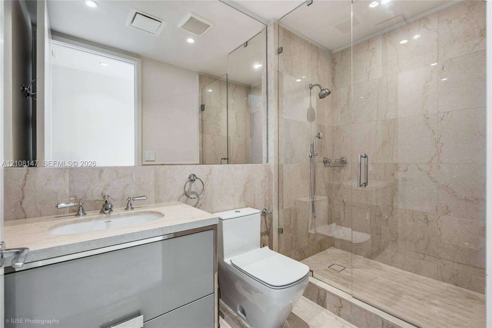 Bathroom featuring a gray vanity with a neutral stone-surfaced countertop, integrated sink, wall-spanning mirror, and a walk-in shower enclosed by a seamless glass door with polished chrome fixtures