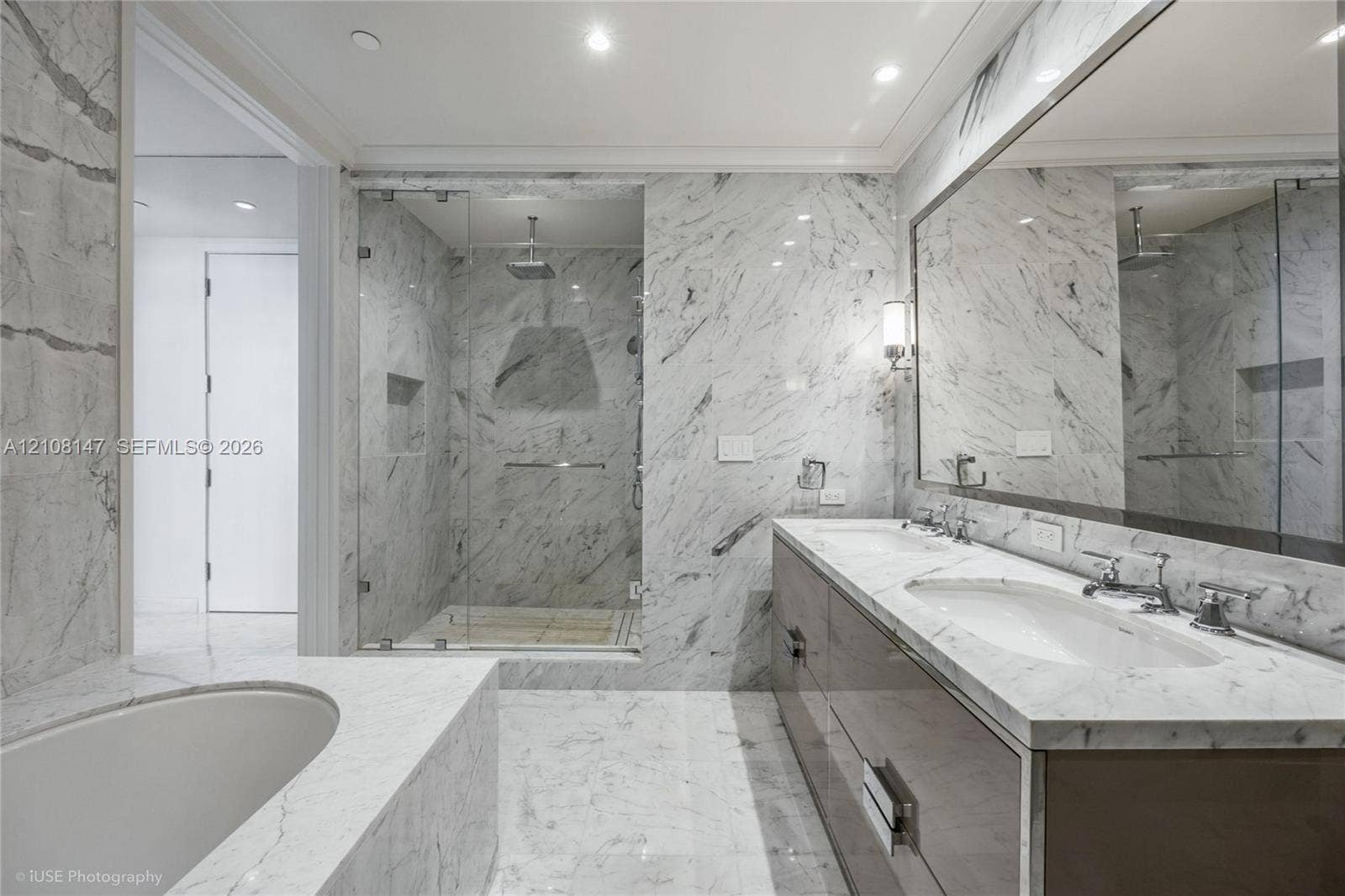 Bathroom featuring extensive stone-surround, frameless glass-enclosed shower with built-in recessed shelving, integrated soaking tub, custom dual-sink vanity with stone countertop, and wall-to-wall mirror