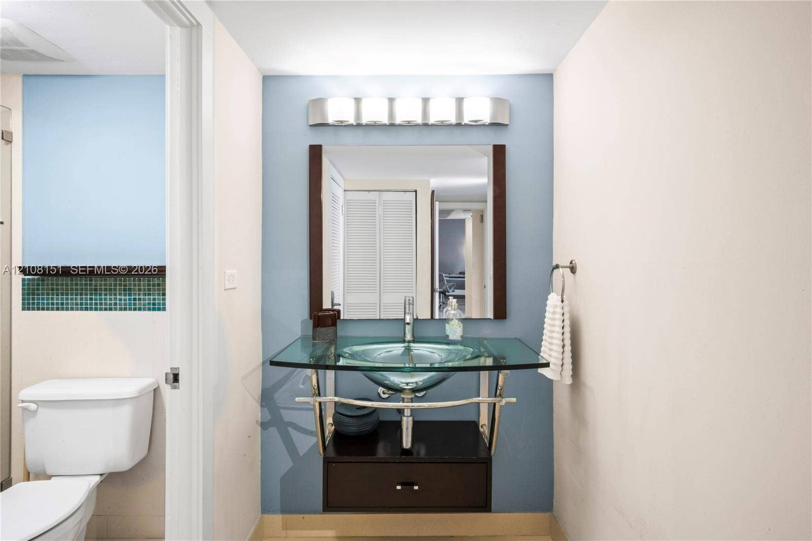 Wall-mounted glass vessel sink, chrome-finished metal console vanity with integrated storage drawer, framed rectangular mirror, and multi-bulb polished metal light fixture