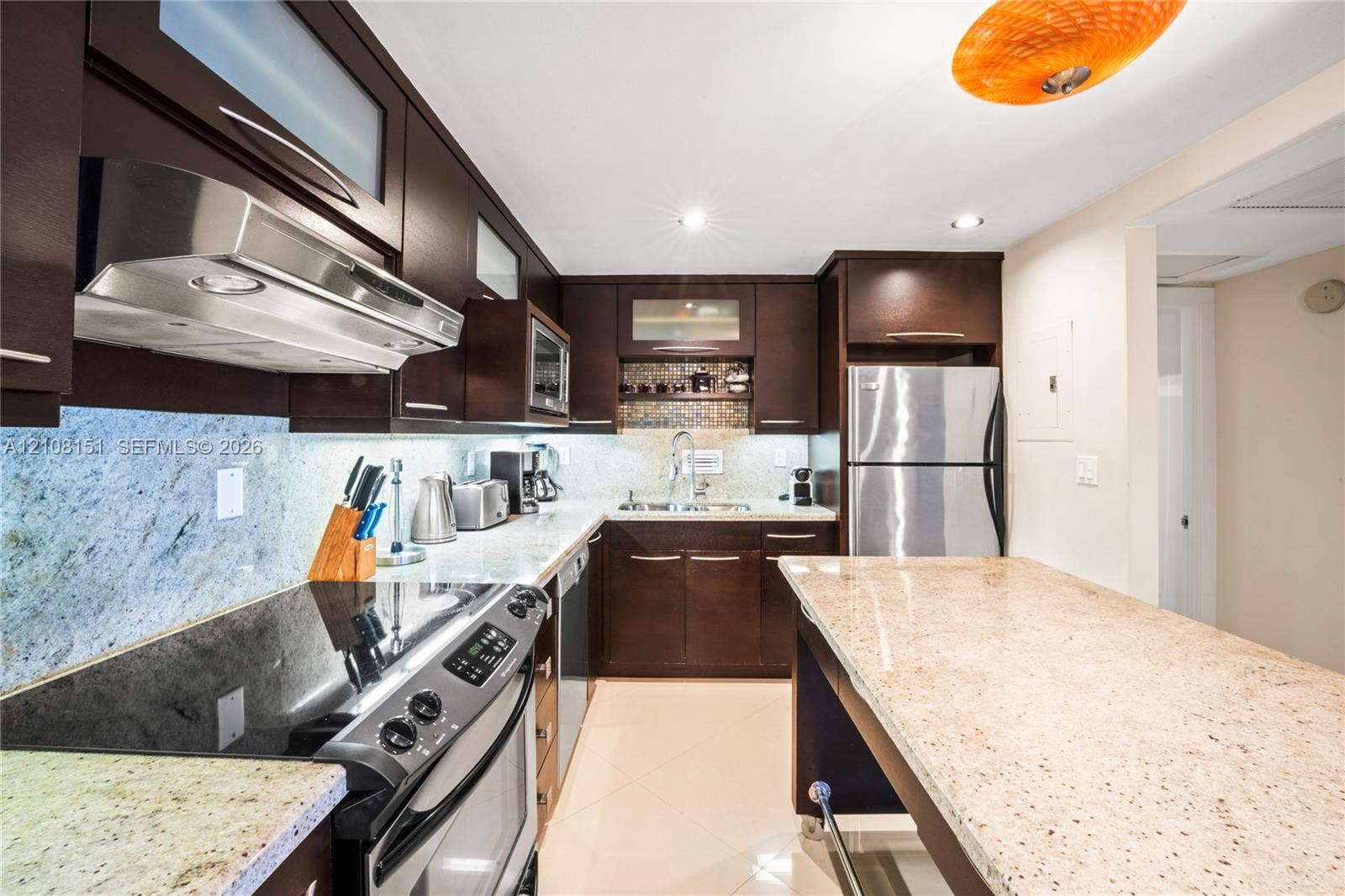 Kitchen featuring dark wood cabinetry, stone countertops, stone tile backsplash, stainless steel appliances, and a central island with an integrated breakfast bar