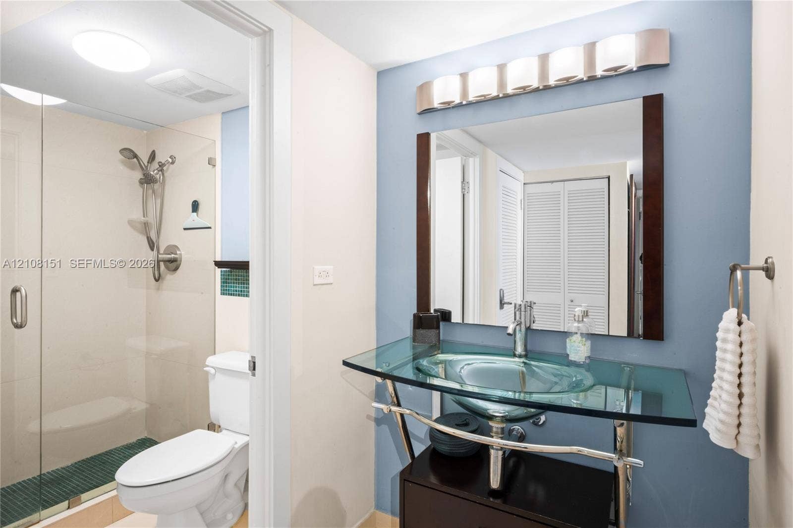 Glass pedestal vanity with integrated basin, wall-mounted multi-light fixture, glass-enclosed shower with handheld showerhead, and framed rectangular wall mirror