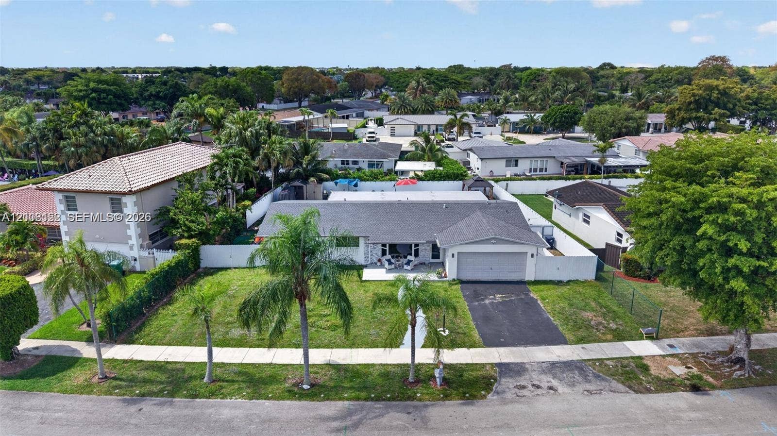 Single-story residence featuring a gray shingled roof, attached one-car garage, white perimeter fencing, and mature palm trees