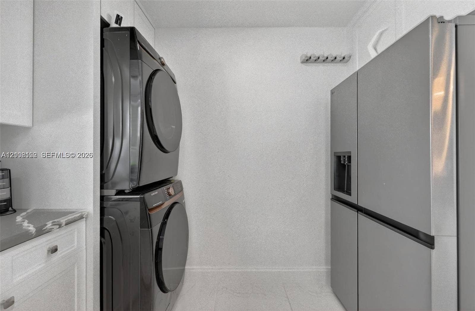 Utility space featuring white cabinetry, a neutral stone-finish countertop, stainless steel refrigerator, and stacked laundry appliances