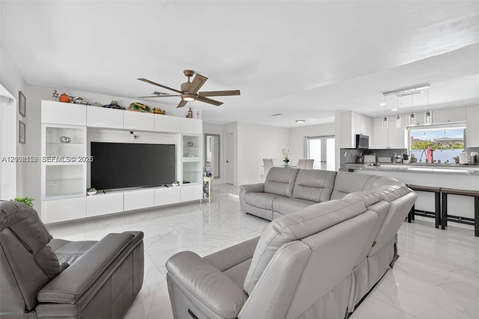 Open-concept interior featuring large-format polished tile flooring, white built-in media cabinetry, a modern ceiling fan, and a breakfast bar