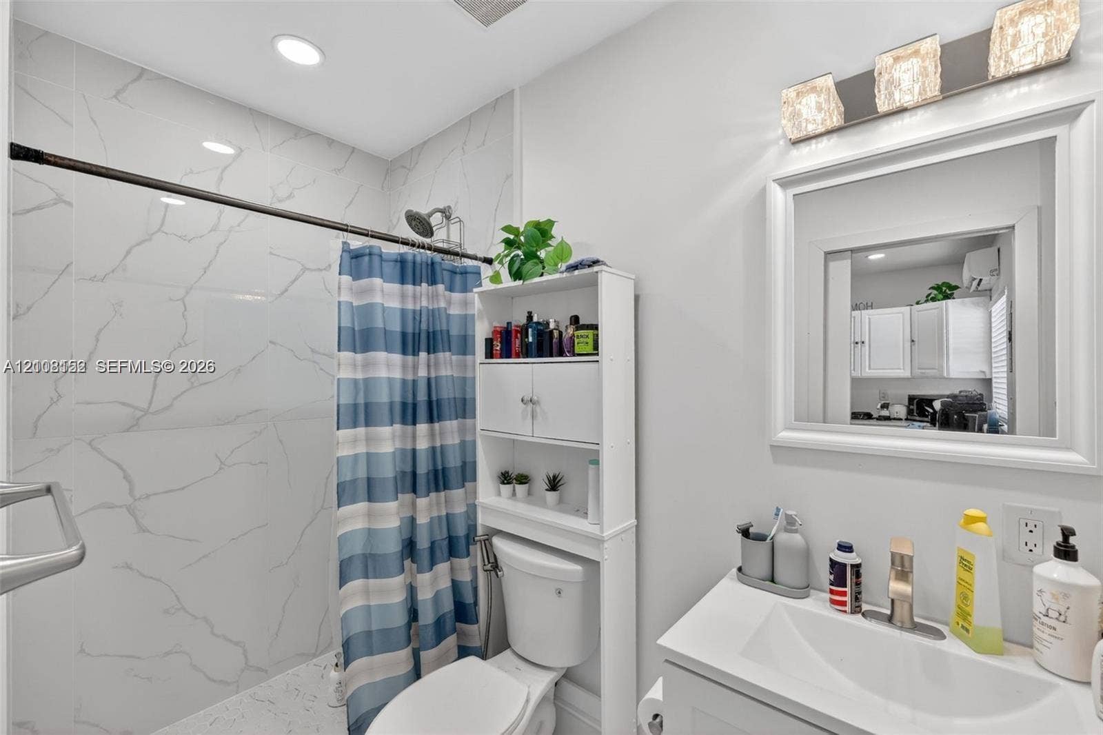 Full-height wall tiling with a white stone-pattern finish, recessed ceiling lighting, three-light decorative wall sconce, framed vanity mirror, and single-basin vanity with a metallic faucet