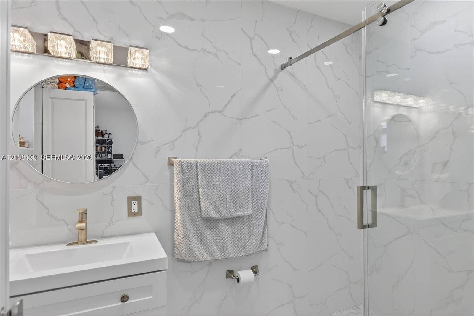 Bathroom featuring large-format wall tiling with veining patterns, a vanity with an integrated rectangular sink, a circular mirror, a glass-enclosed shower with metal hardware, and a four-light fixture