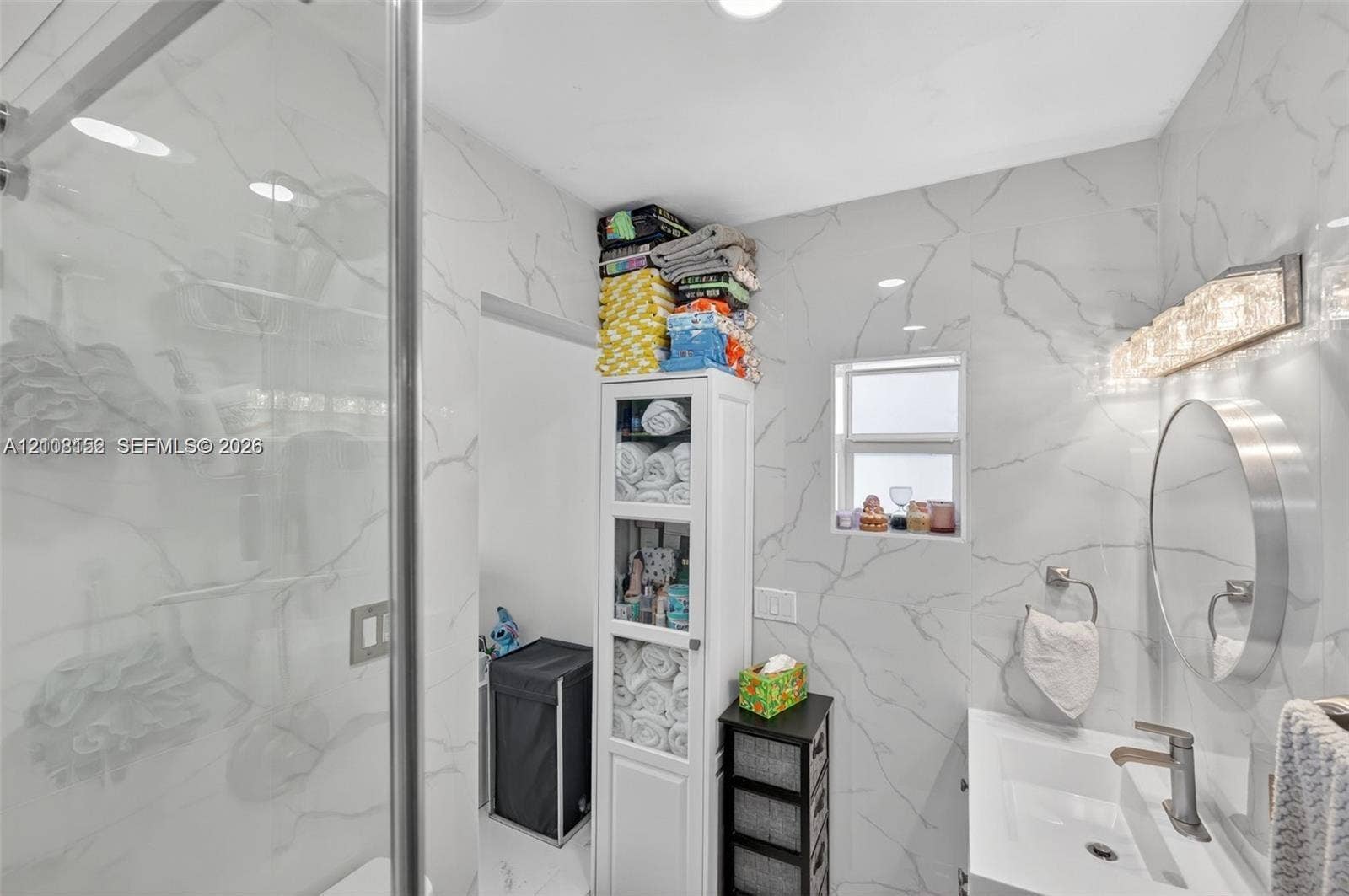 Full-height, large-format stone-patterned wall tile, glass-enclosed shower, round wall-mounted mirror, integrated rectangular sink with metallic faucet, and multi-bulb vanity lighting