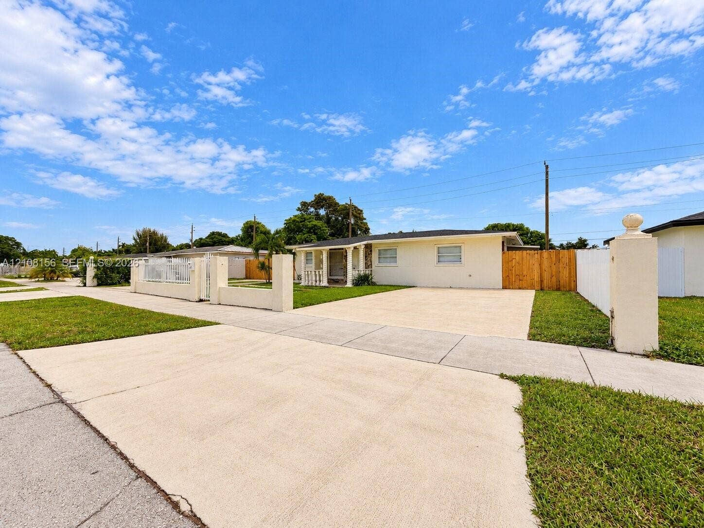 17175 NW 41st Avenue, Miami Gardens, FL 33055 Single-story exterior featuring a light-colored stucco facade, expansive concrete driveway, gated front perimeter wall, and wood-panel privacy fencing