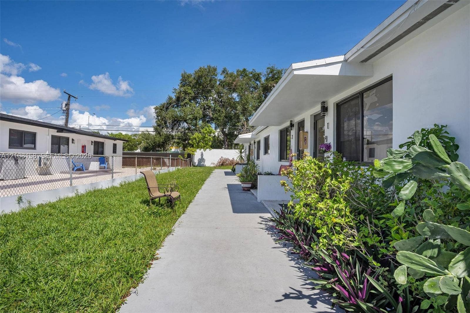 Exterior of building featuring white stucco siding, a concrete walkway, chain-link fencing, and established shrubbery