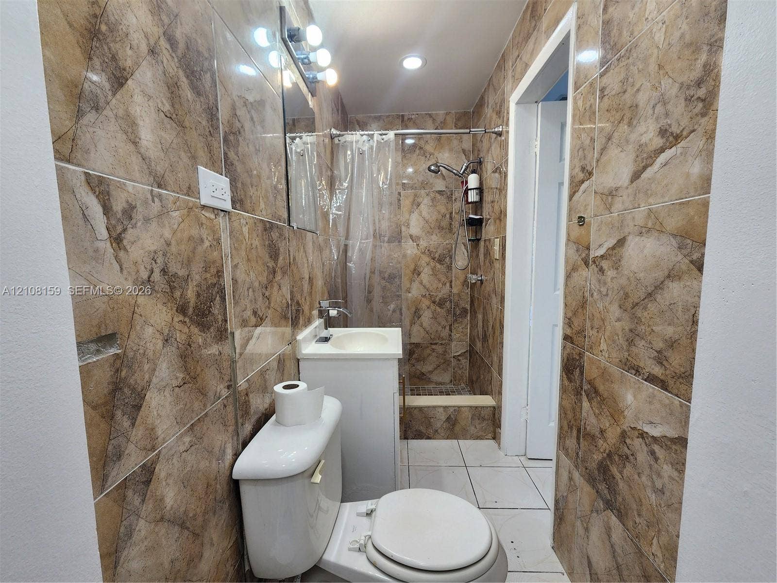 Full-height wall tiling with a decorative stone pattern, white vanity with an integrated sink, walk-in shower with a handheld spray head, and recessed ceiling lighting