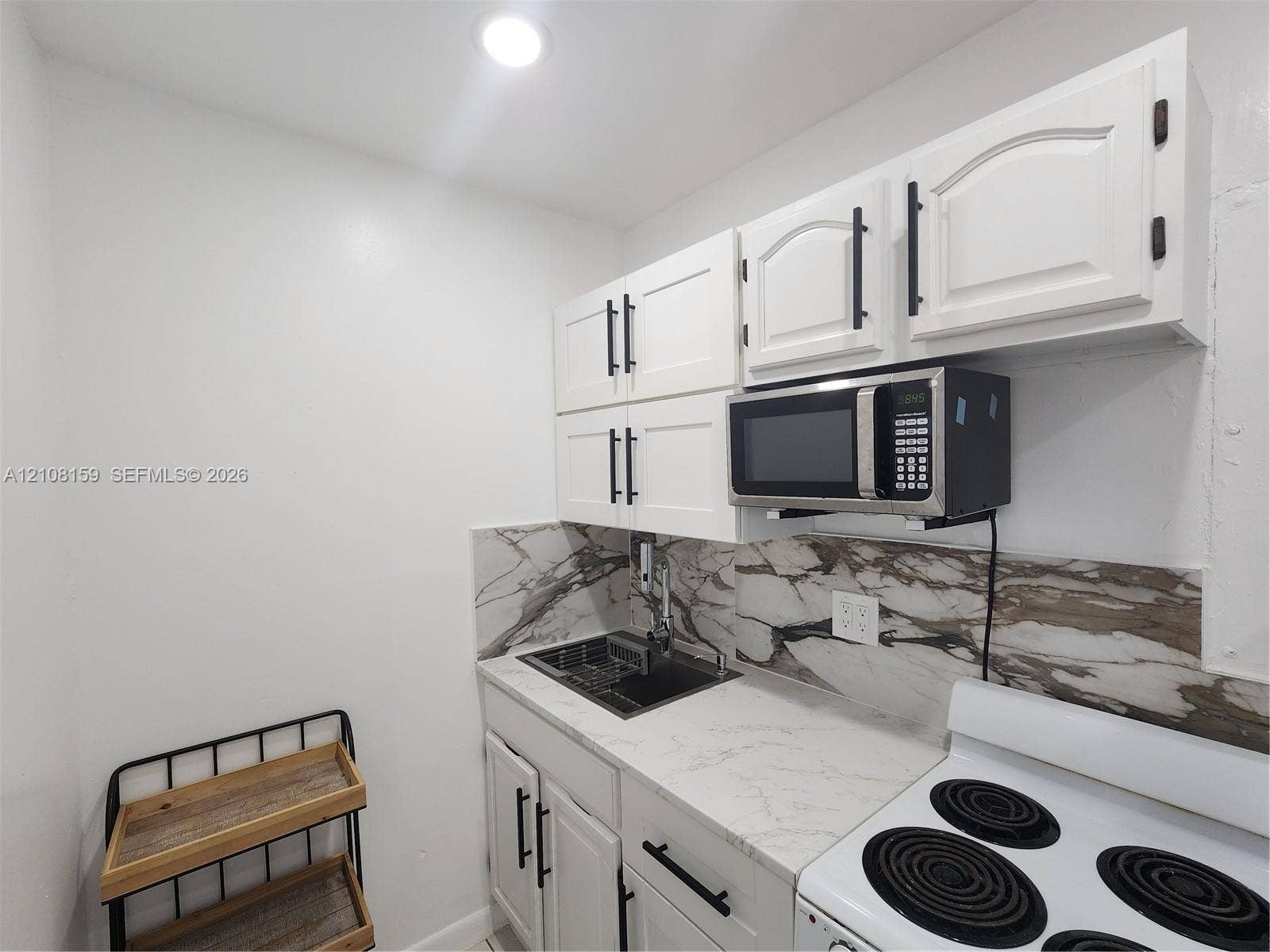 White shaker-style cabinetry with matte black hardware, stone-patterned backsplash, light-toned stone-finish countertop, stainless steel drop-in sink, and integrated microwave