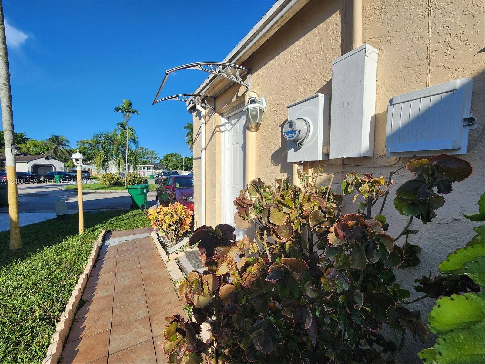 Stucco exterior featuring a tiled walkway, metal door awning, exterior wall-mounted light fixture, and mature landscape plantings