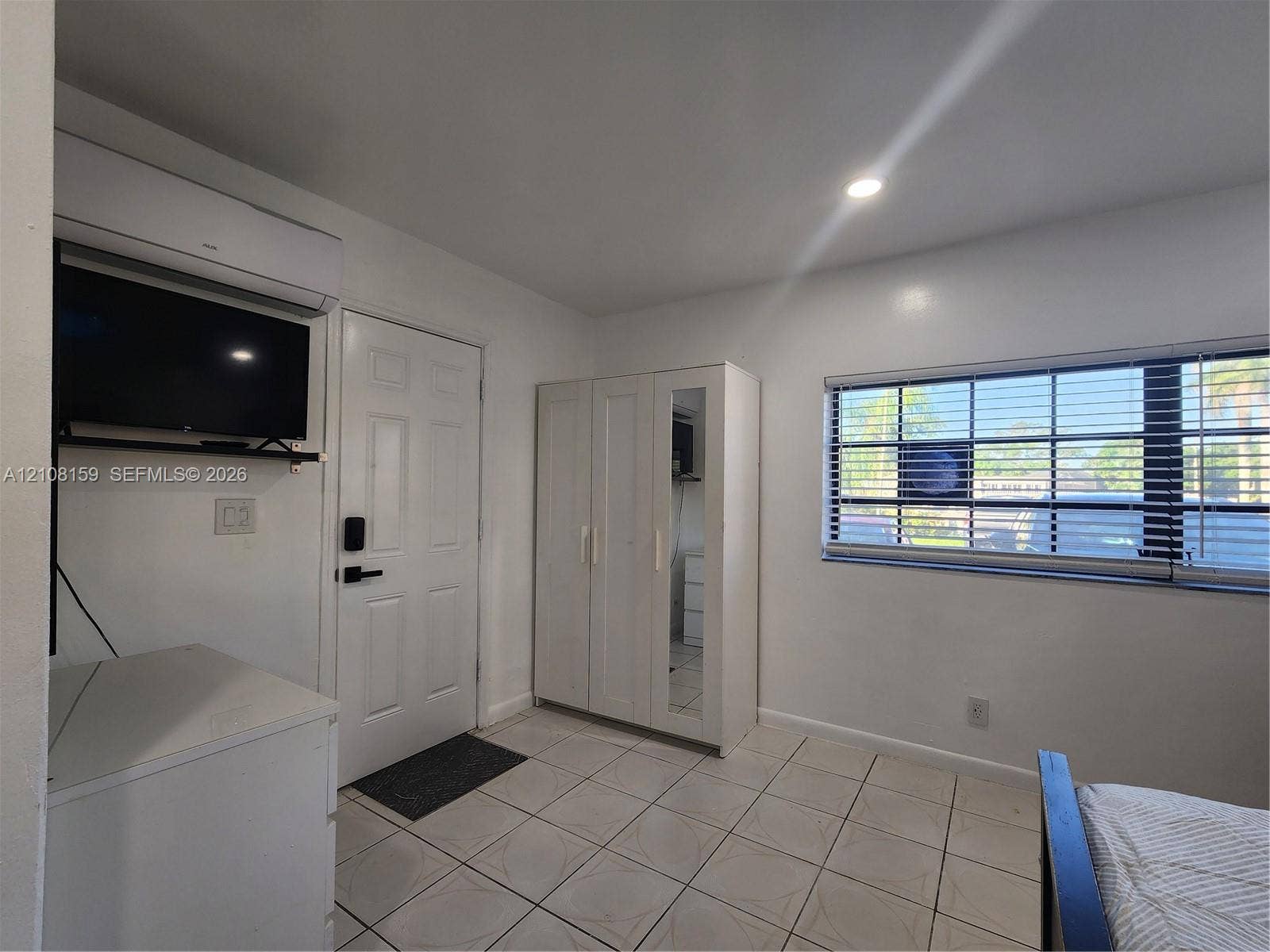 Room featuring neutral tile flooring, a wall-mounted climate control unit, a multi-pane window with horizontal blinds, and a built-in wardrobe with a mirrored door