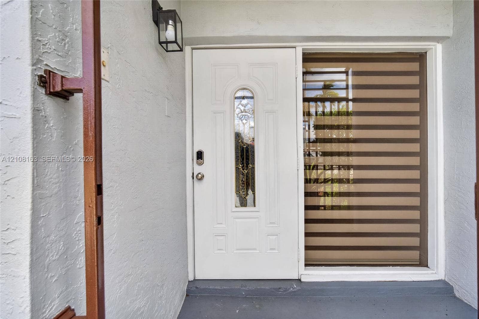 11510 SW 5th Street, #1, Sweetwater, Sweetwater, FL 33174 Entryway featuring a white exterior door with a decorative glass insert, integrated smart lock, metal wall-mounted lantern, and sidelight window with horizontal banded privacy shades