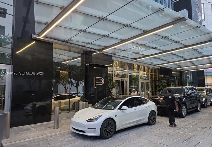 Covered porte-cochre featuring a glass canopy with integrated linear lighting, polished black stone facade, and stone-paved motor court