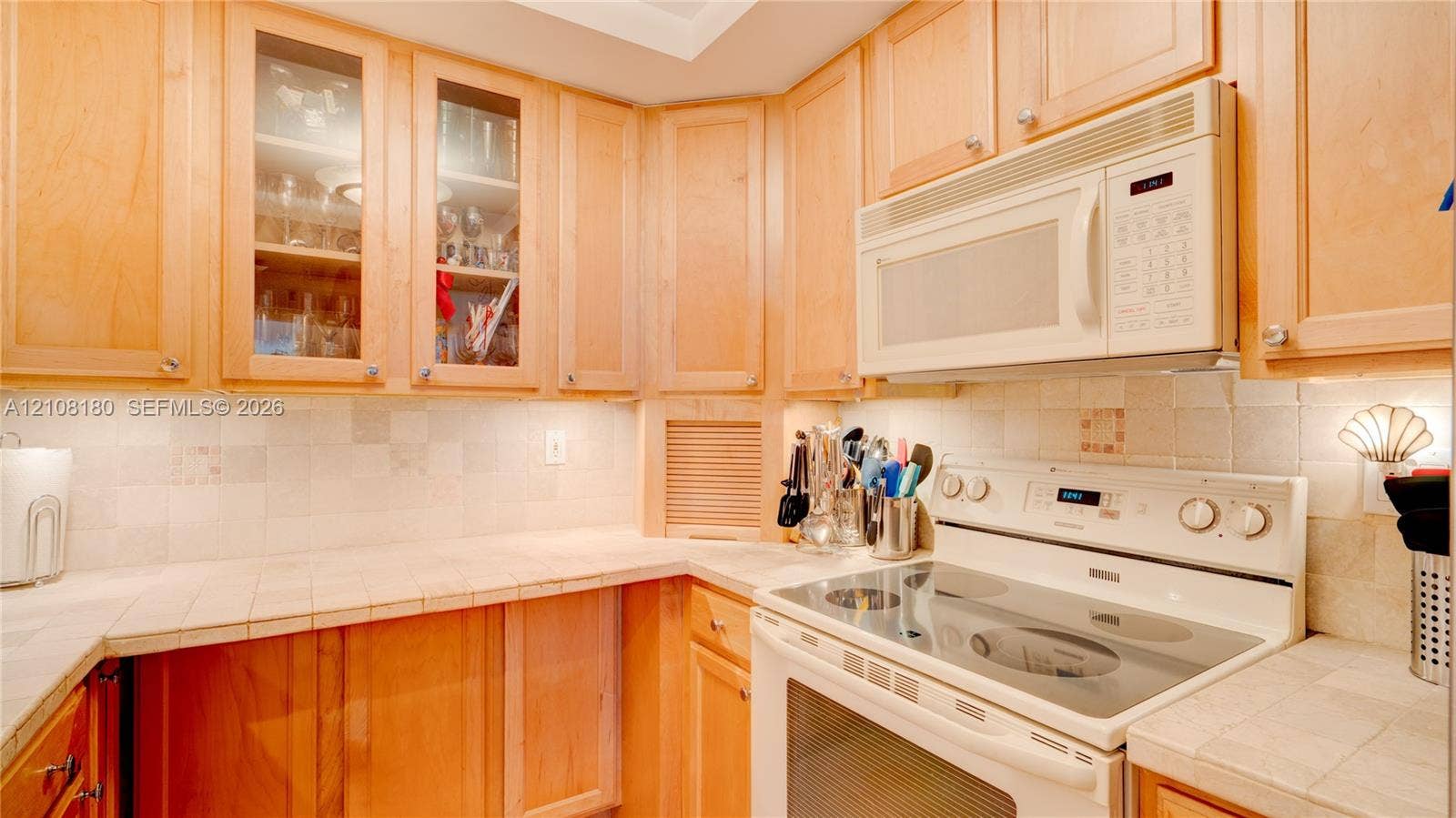 2457 Collins Avenue, #1501, Miami Beach, FL 33140 Kitchen featuring natural wood cabinetry, glass-front upper display cabinets, tile backsplash, tile countertops, and integrated over-the-range microwave