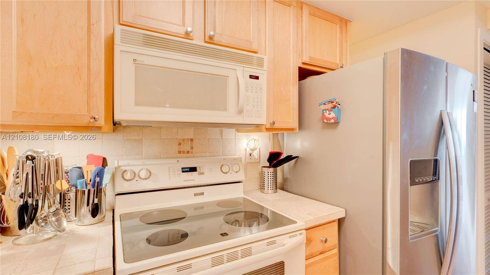 2457 Collins Avenue, #1501, Miami Beach, FL 33140 Light wood-finish cabinetry, white tile backsplash, integrated over-the-range microwave, smooth-top electric range, and stainless steel refrigerator