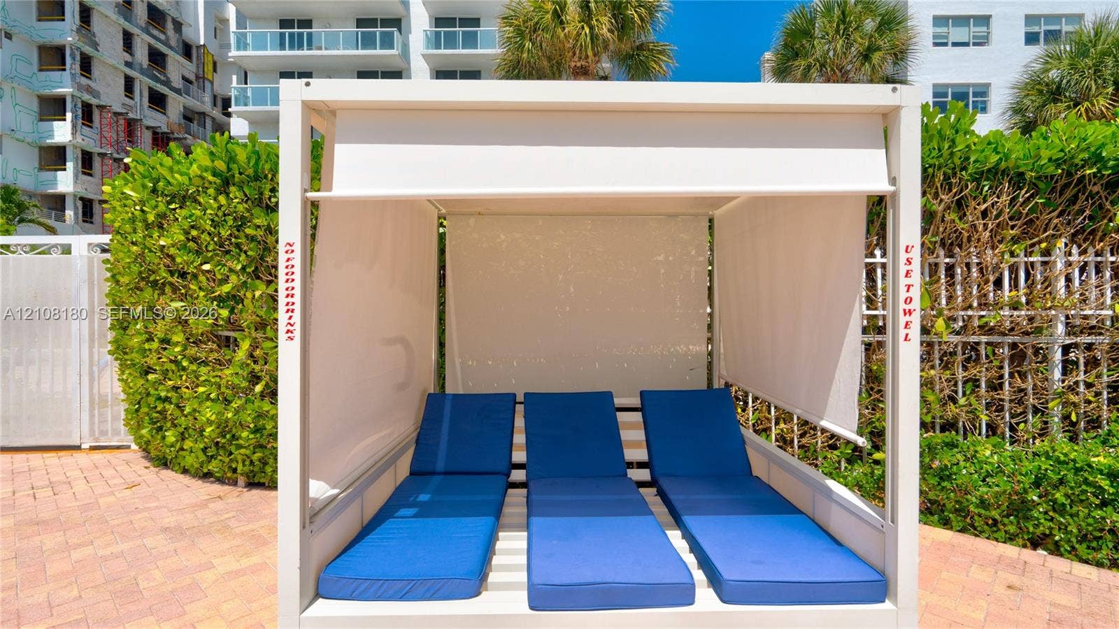 2457 Collins Avenue, #1501, Miami Beach, FL 33140 Permanent poolside cabana featuring a white metal frame, integrated retractable fabric shades, and tiered cushioned platforms situated on a brick paver deck