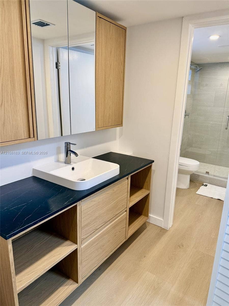 101 Ocean Lane Drive, #205, Key Biscayne, Key Biscayne, FL 33149 Wood-finish vanity featuring open shelving, a dark countertop, integrated vessel sink, and mirrored medicine cabinets, with wood-finish flooring extending into the tiled shower area