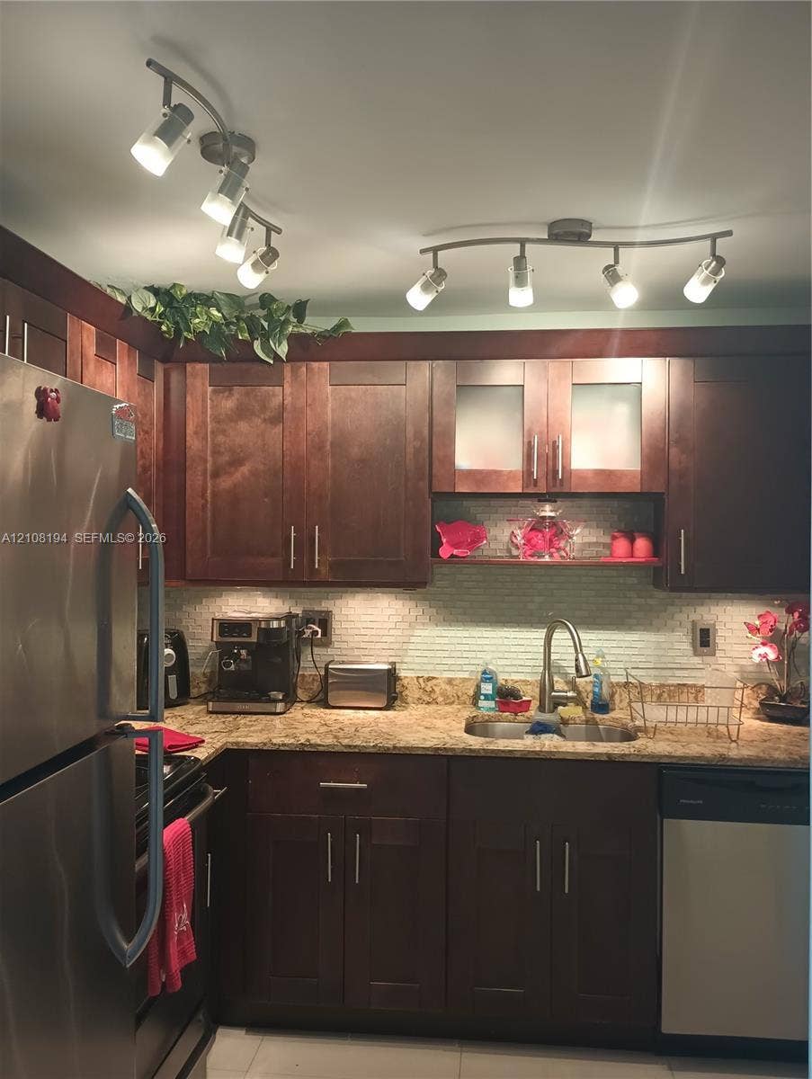 Dark wood cabinetry featuring brushed metal hardware, stone-finish countertops, textured tile backsplash, stainless steel appliances, and dual adjustable track lighting fixtures