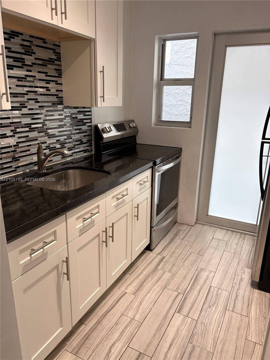 Kitchen featuring white cabinetry with brushed metal hardware, dark stone-finish countertops, stainless steel undermount sink, multi-tonal mosaic tile backsplash, and wood-finish flooring