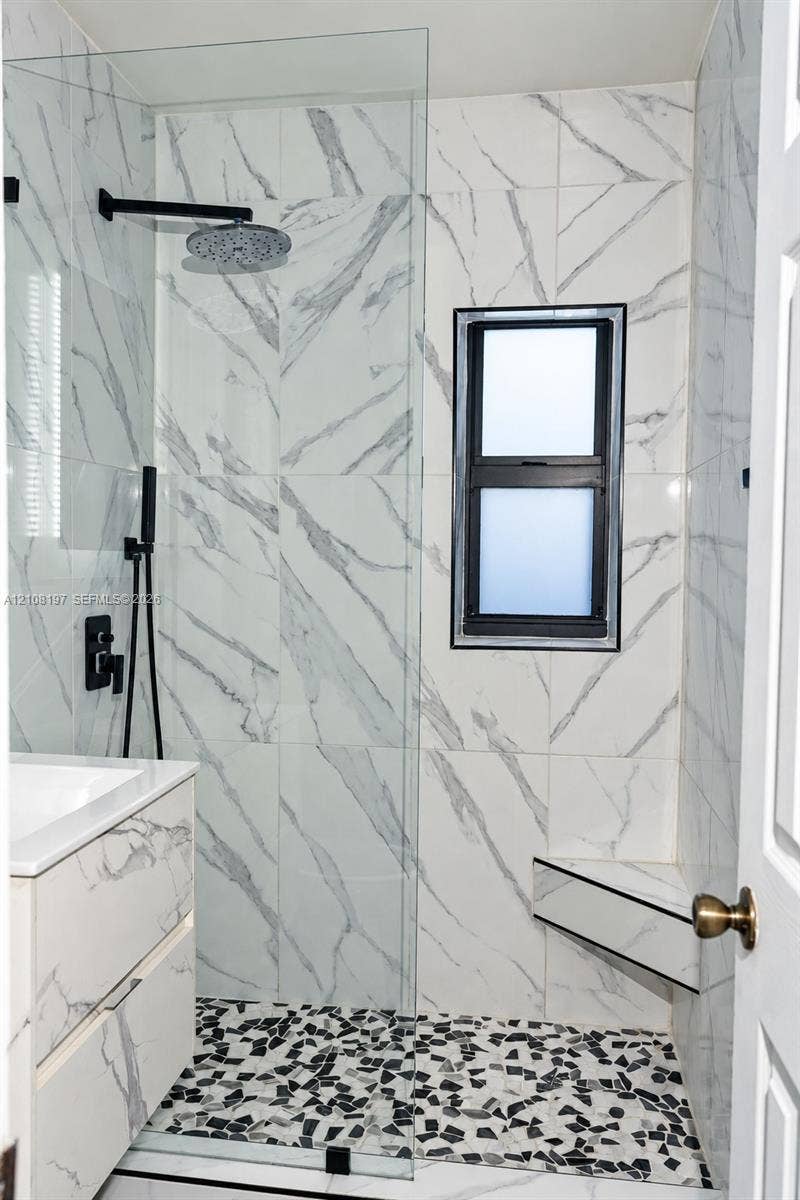 Walk-in shower featuring large-format stone-patterned wall tile, a built-in corner seat, matte black rainfall showerhead with handheld attachment, pebble-texture flooring, and a frameless glass partition