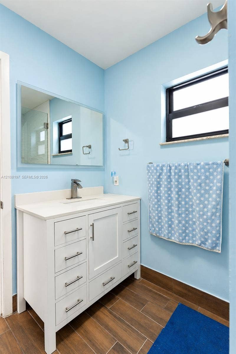 White vanity featuring multiple storage drawers, rectangular basin, and single-lever faucet, complemented by a frameless wall-mounted mirror, wood-finish tile flooring, and metal hardware