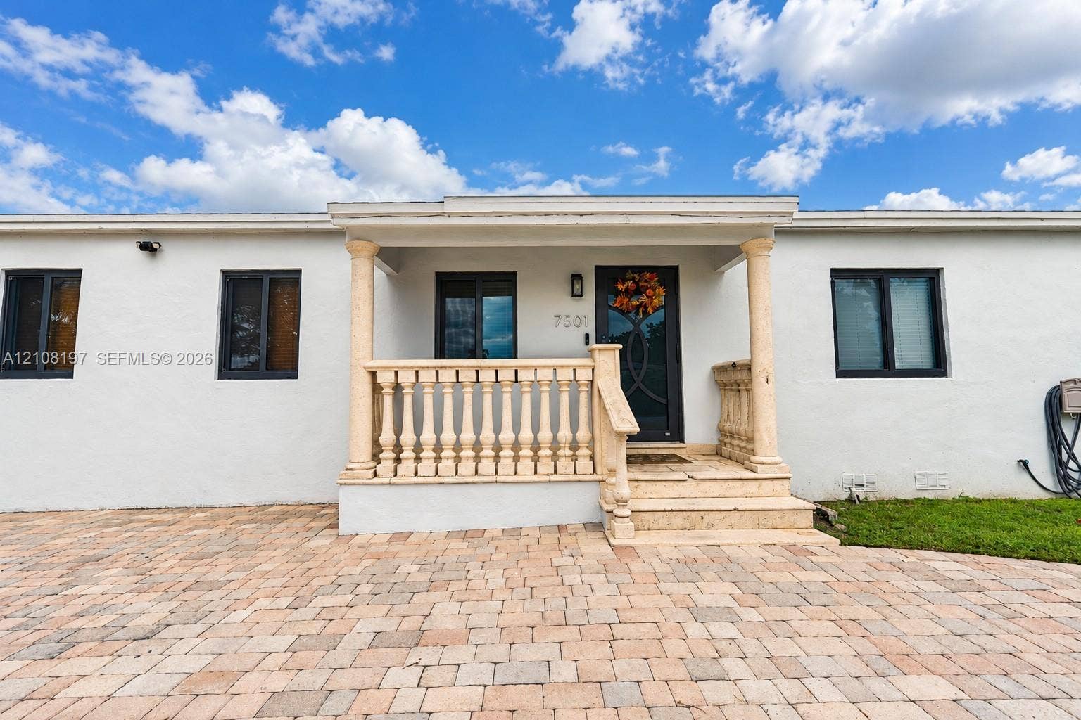 Stucco exterior featuring a covered front porch with stone-finish balustrade, columns, and steps, complemented by an expansive interlocking paver driveway and dark-frame windows