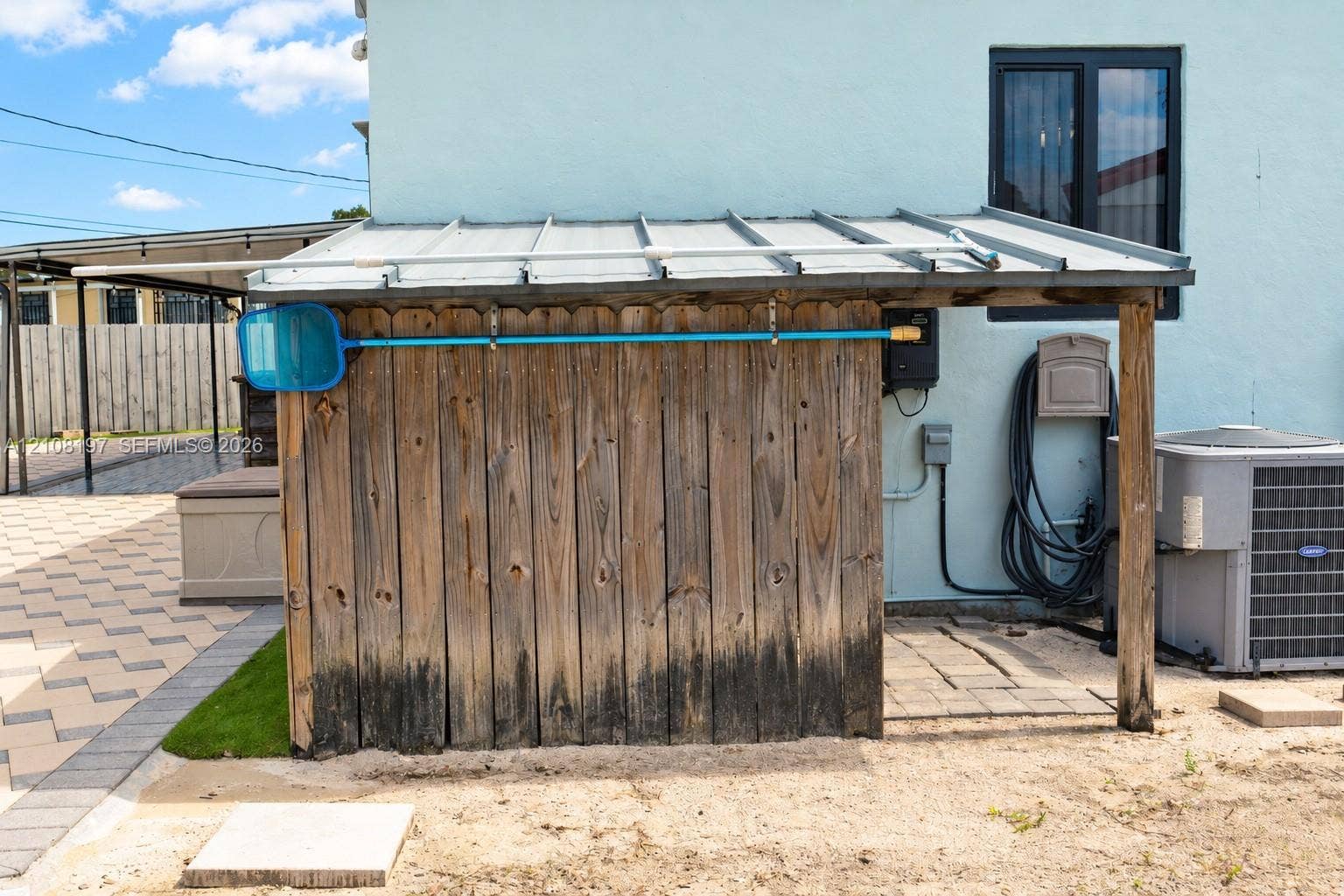 Exterior storage structure with a metal roof and wood vertical planking, featuring an open-sided design, stone paver flooring, and a dedicated central air conditioning unit