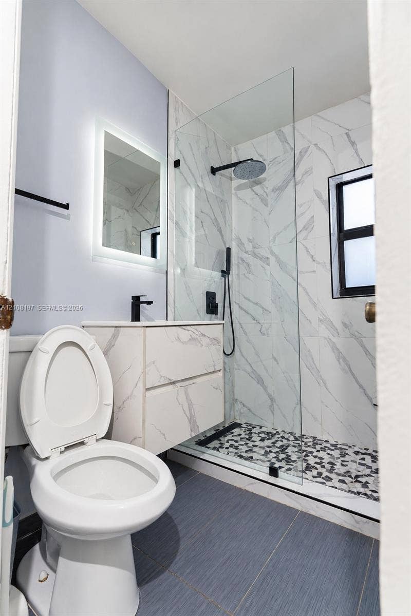 Floating vanity with stone-finish cabinetry, walk-in shower with frameless glass enclosure, rain-style showerhead, and wall-mounted backlit mirror