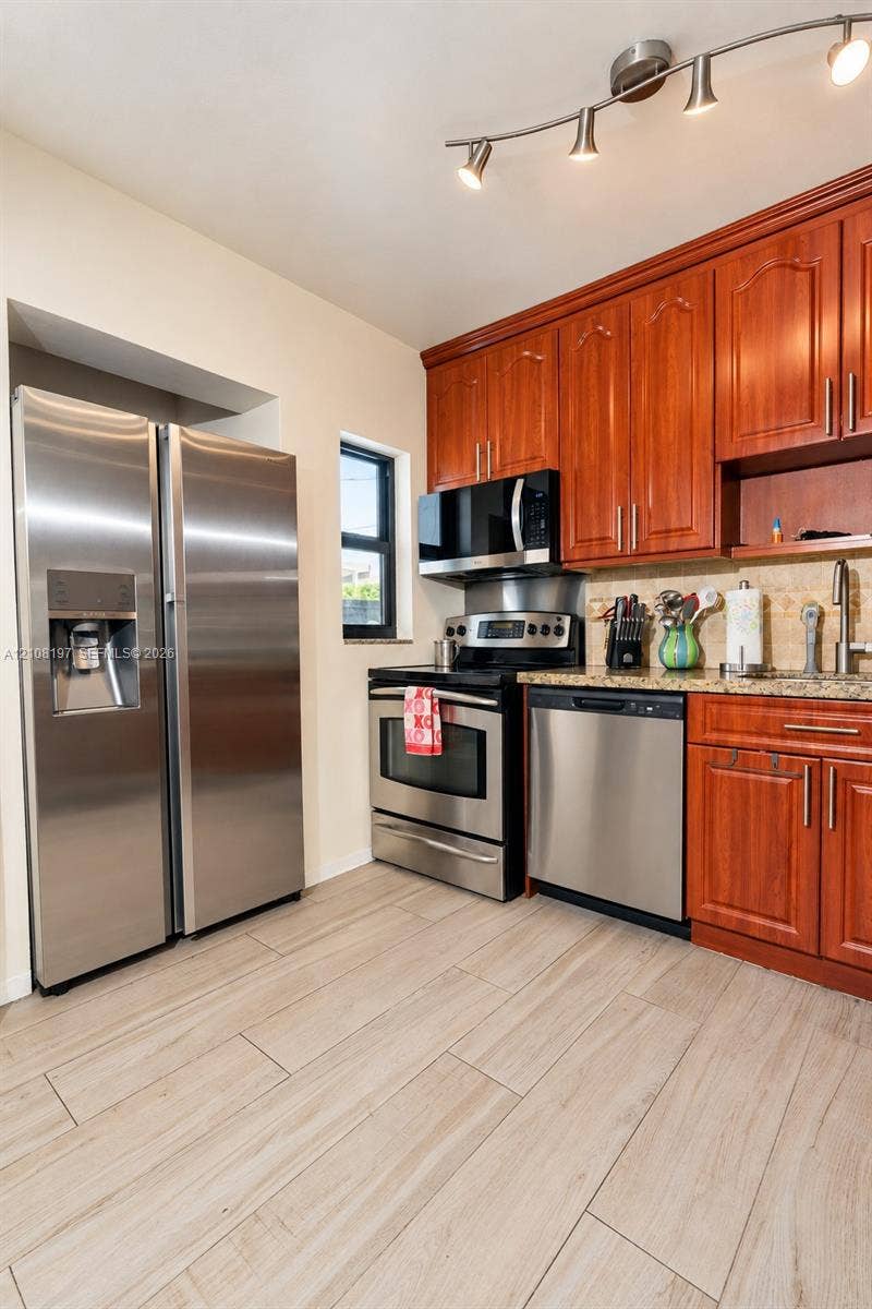 Kitchen featuring wood-finish cabinetry, stainless steel appliances, stone-surface countertops, tile backsplash, and wood-finish tile flooring