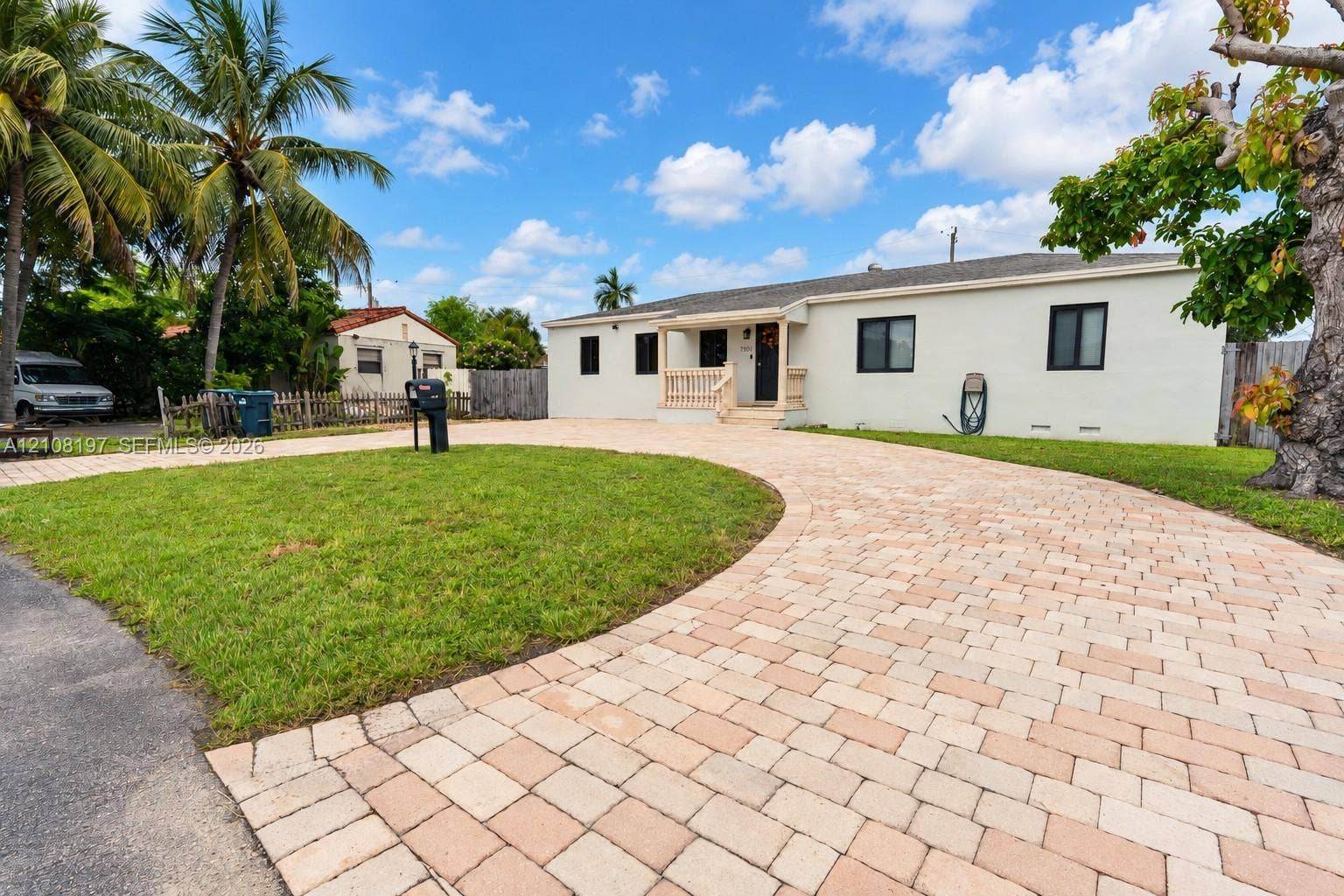 Single-story residence featuring light-toned exterior finish, a sprawling paver driveway, manicured lawn, and mature palm trees