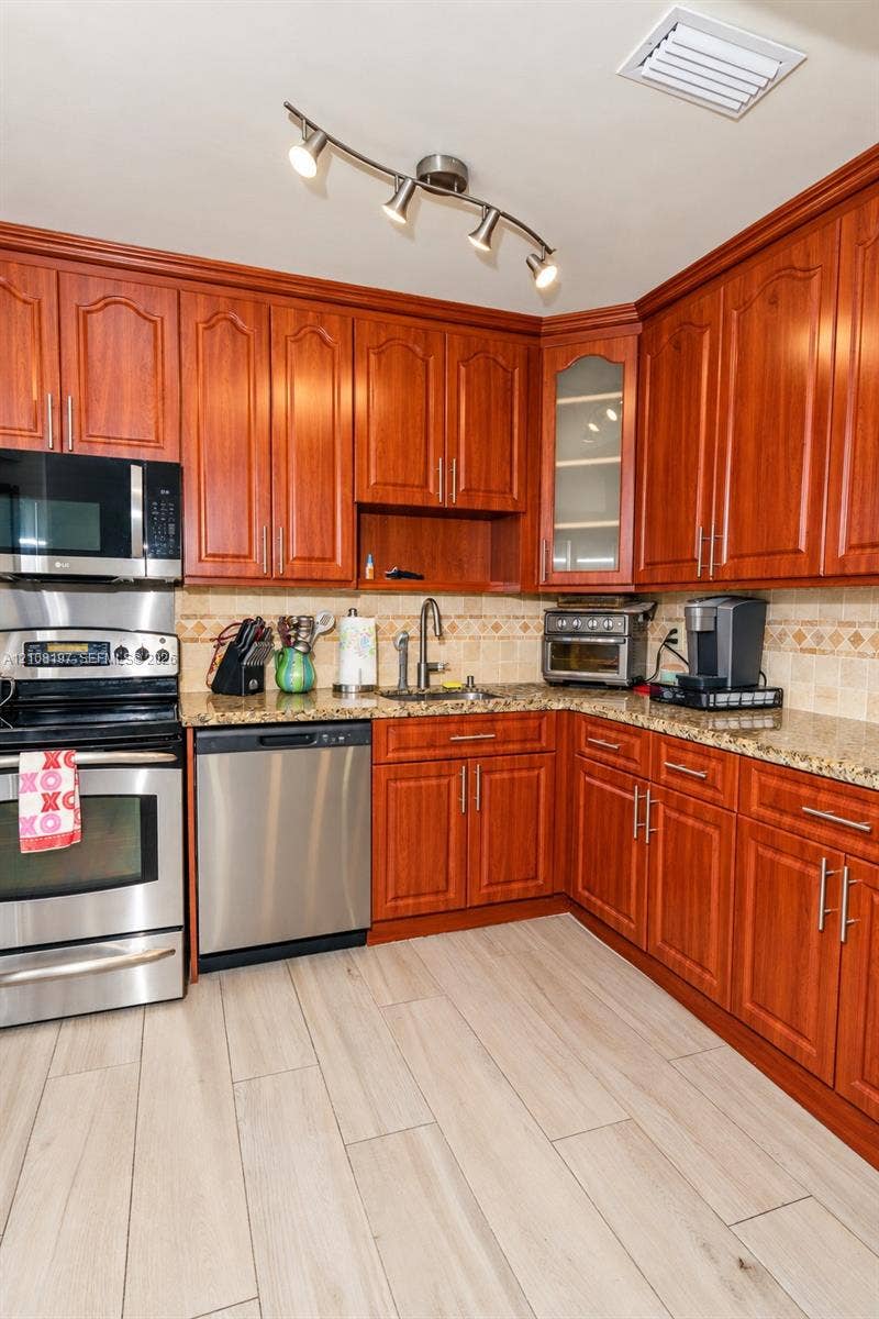 Kitchen featuring raised-panel cherry wood cabinetry, speckled stone countertops, stainless steel appliances, decorative tile backsplash, and wood-finish flooring
