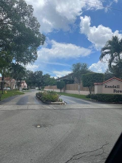 Community entrance featuring masonry signage, mature trees, manicured hedges, and palm landscaping