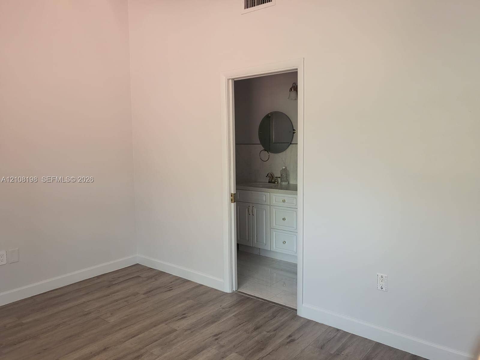 Room featuring wood-finish flooring and a doorway leading to a bathroom equipped with a white vanity cabinet, light-colored countertop, and a circular wall-mounted mirror
