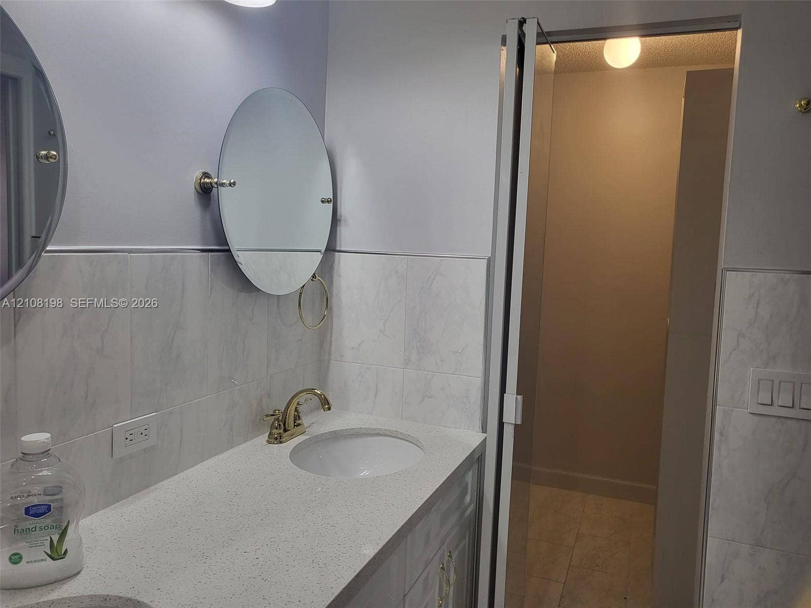 Bathroom vanity featuring a speckled countertop, integrated undermount sink, brass-finish faucet, and oval wall-mounted mirrors, complemented by stone-tile wainscoting and a glass-enclosed shower