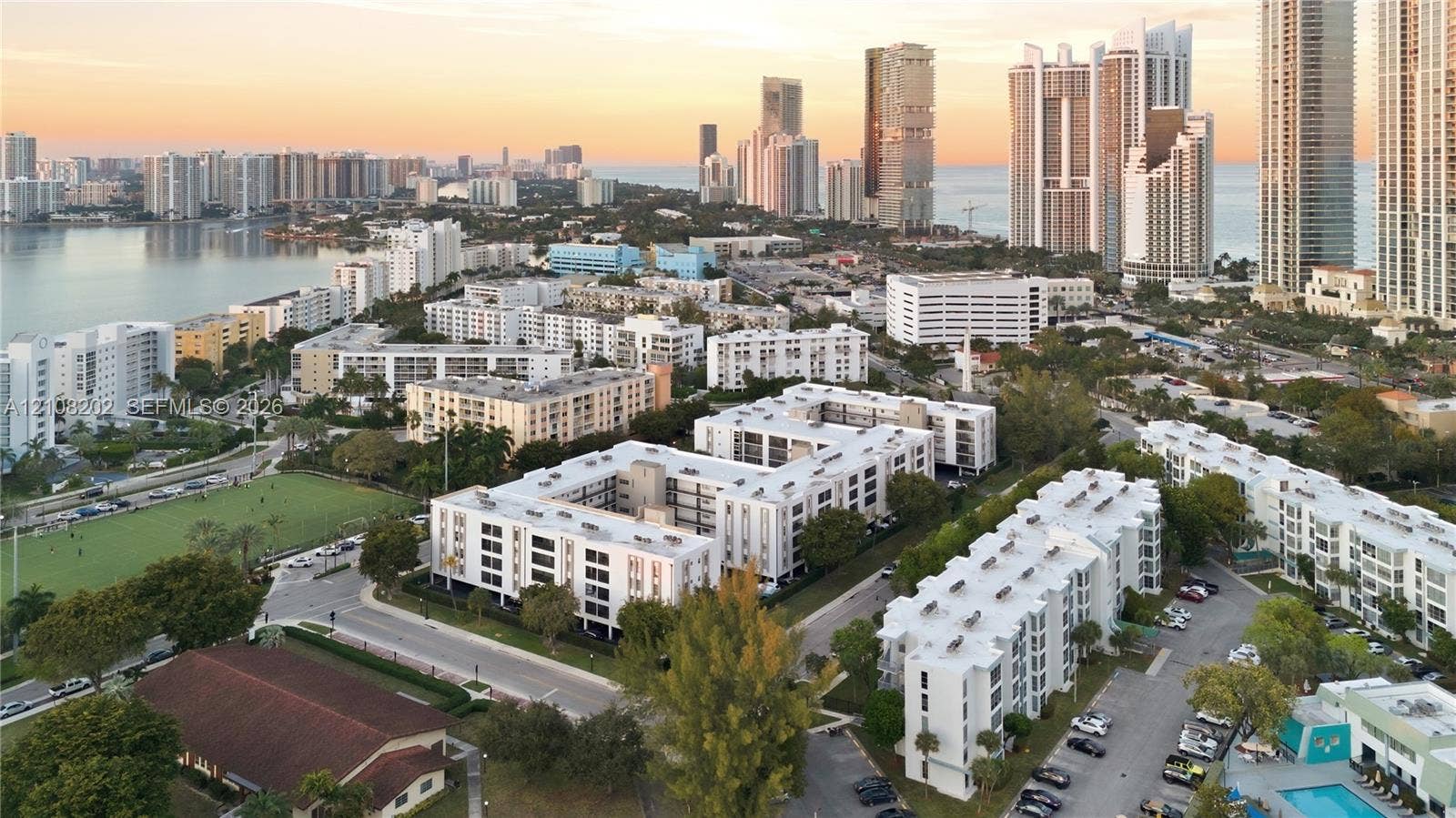 Aerial perspective of the property grounds featuring multi-story residential buildings, paved parking areas, mature landscaping, and proximity to coastal waters and urban skyline