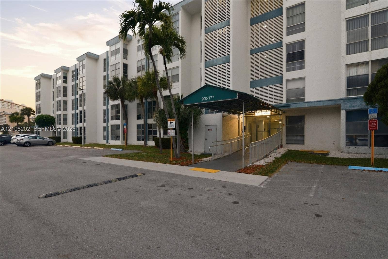Multi-story exterior featuring a concrete facade, covered entryway with a metal awning, paved surface parking, and mature palm trees