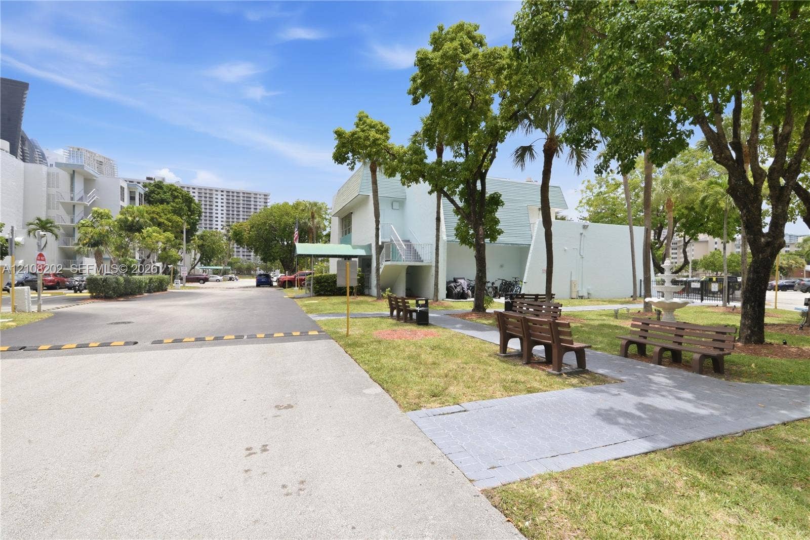Exterior grounds featuring paved walkways, manicured lawn, mature trees, and fixed-position park benches