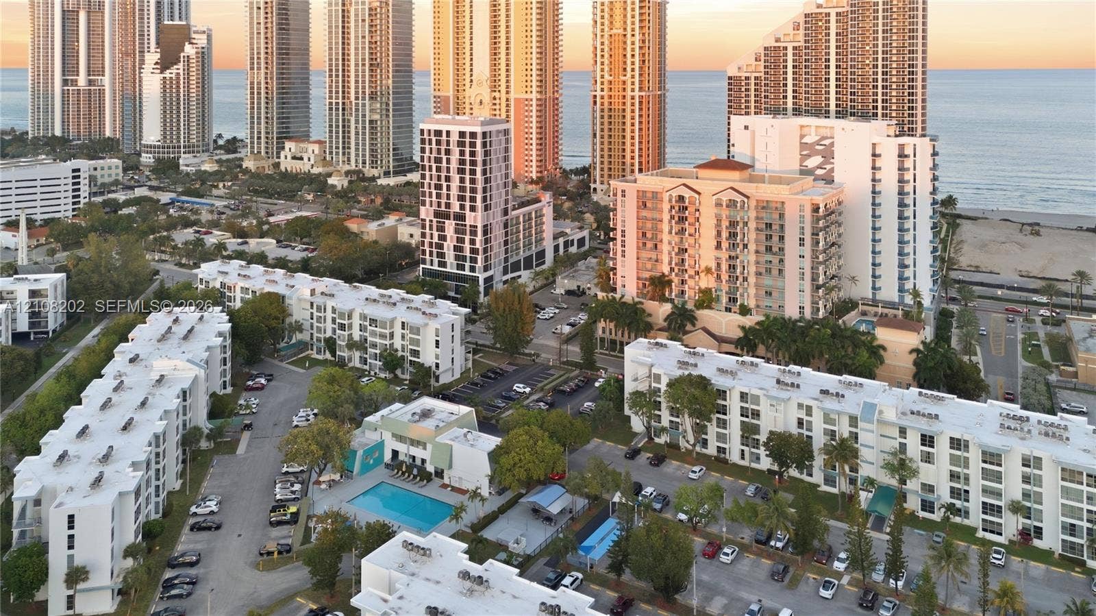 Aerial perspective of a multi-building residential complex featuring a central swimming pool, paved surface parking areas, and mature tropical landscaping, situated near the coastline
