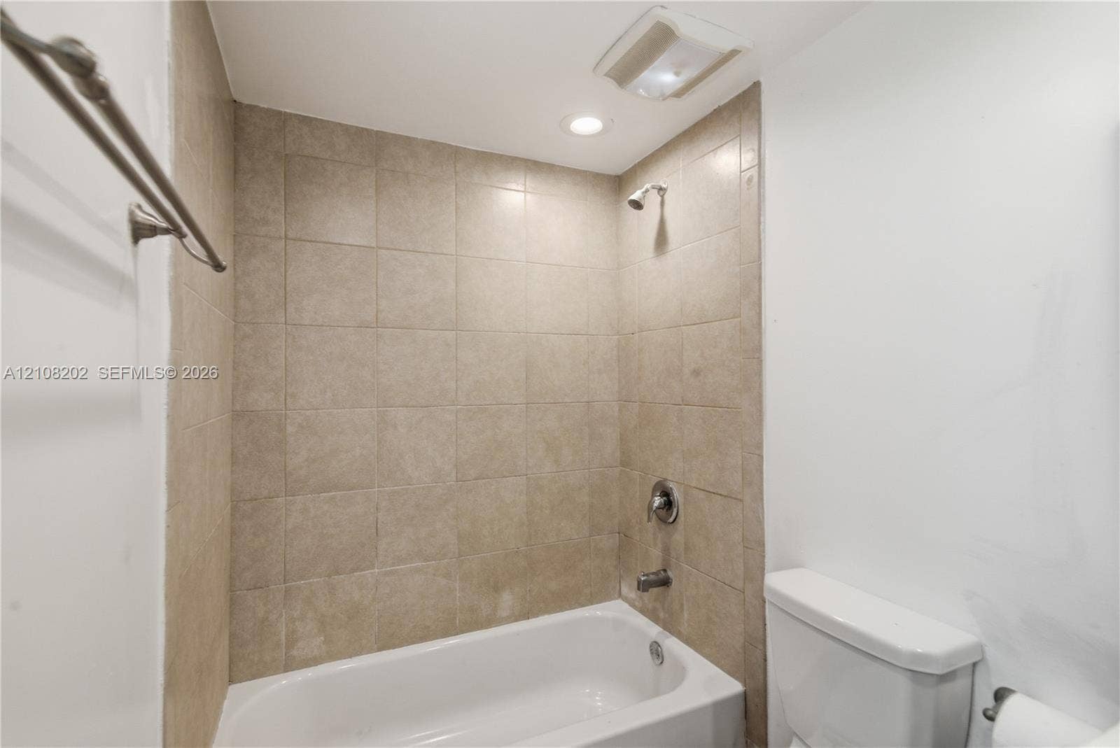 Bathtub with surround of neutral-toned square ceramic tiles, integrated chrome shower fixture, and recessed lighting