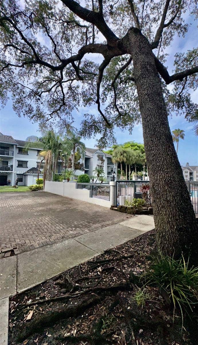 Coral Springs, Coral Springs, FL 33071 Exterior grounds featuring paved brick surfacing, mature canopy trees, concrete walkways, and metal perimeter fencing