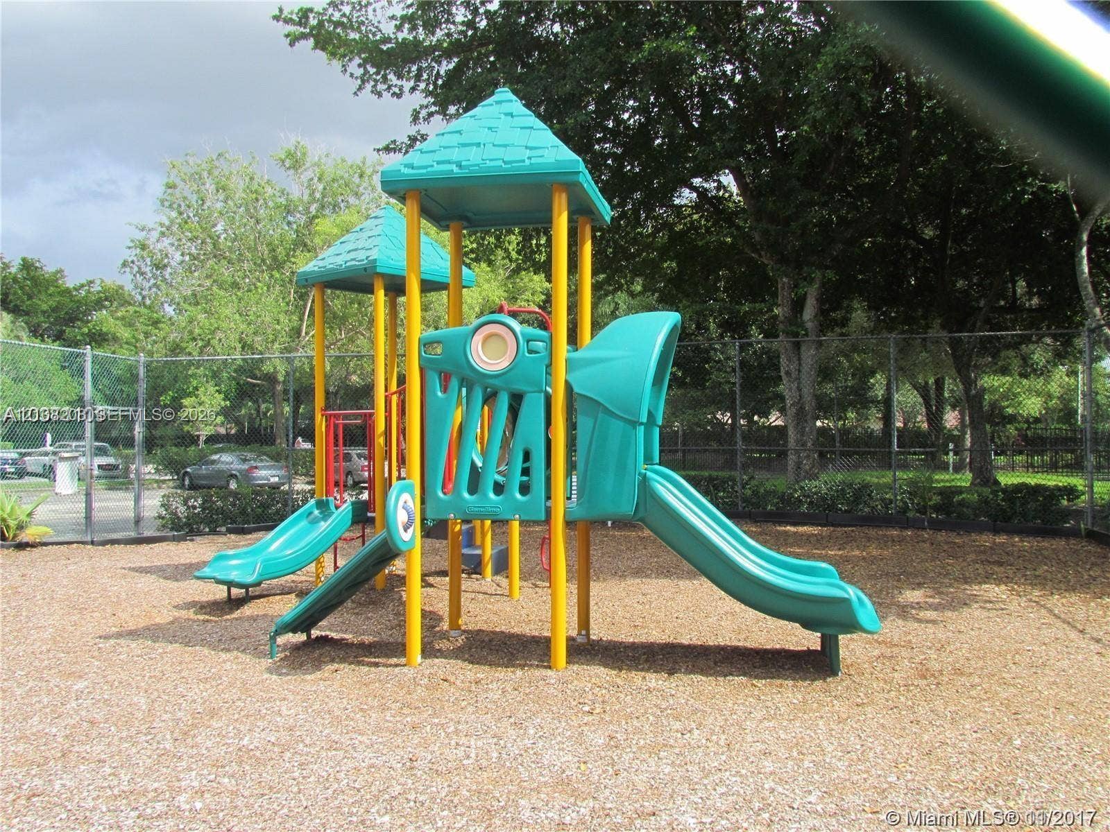 Coral Springs, Coral Springs, FL 33071 Outdoor recreational structure featuring a multi-level composite play system, dual slides, and wood-chip ground covering, enclosed by chain-link fencing