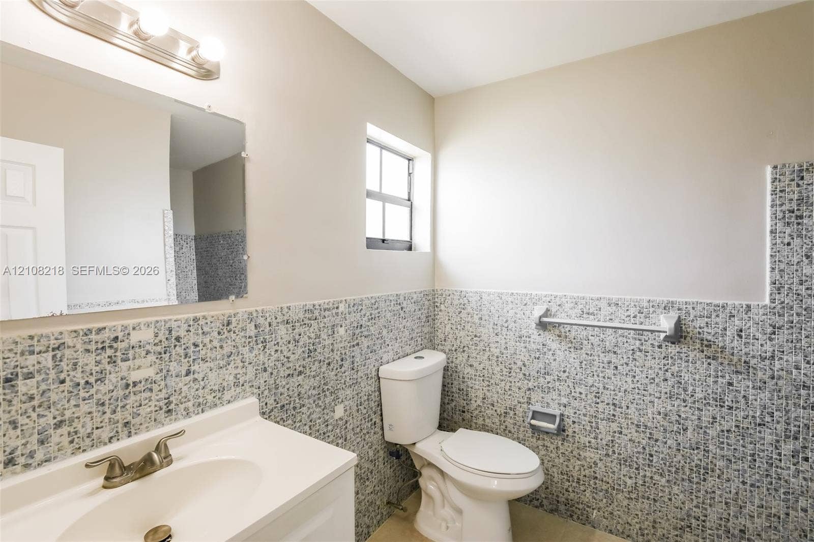 Bathroom featuring mosaic tile wainscoting, a vanity with a white countertop, wall-mounted metallic light fixture, and a window