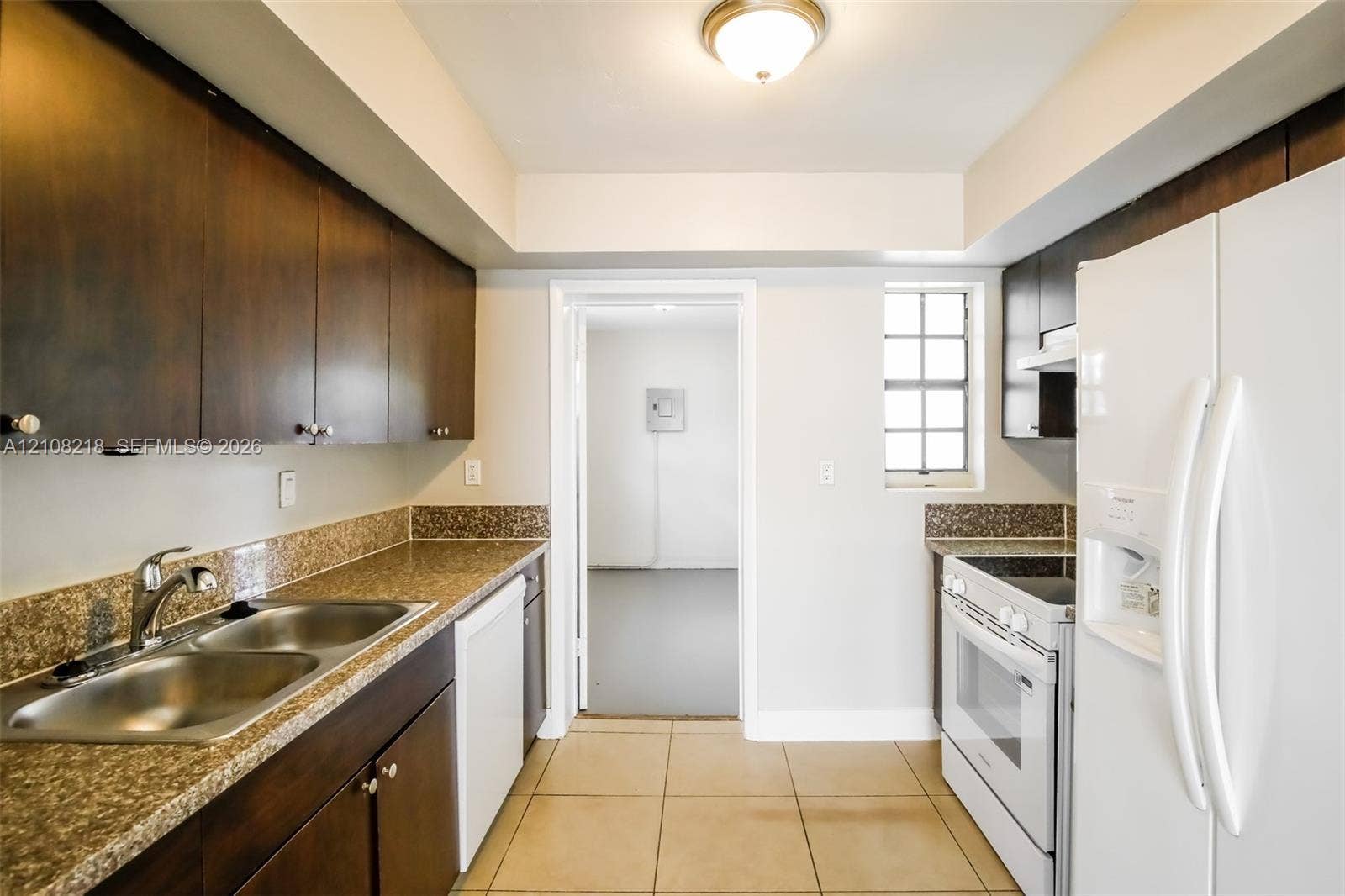 Kitchen featuring dark wood-finish cabinetry, stone-surface countertops, double-basin stainless steel sink, ceramic tile flooring, and integrated white appliances