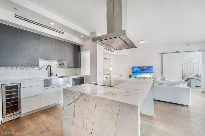 Kitchen featuring a waterfall-edge island with stone surfacing, stainless steel farmhouse sink, matte finish upper cabinetry, integrated wine refrigerator, and stainless steel overhead range hood