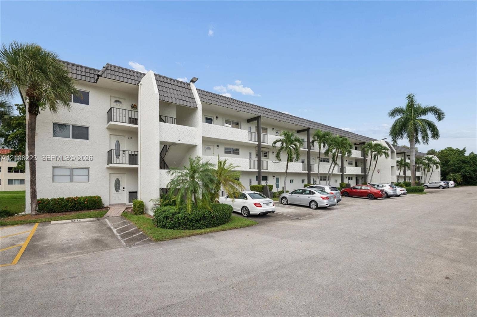 311 S Hollybrook Drive, #309, Pembroke Pines, FL 33025 Multi-story stucco building exterior featuring a textured tile roof, private exterior balconies, and mature palm landscaping